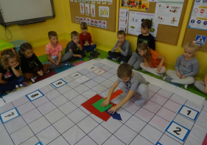 Grupa dzieci siedzi wokół rozłożonej na dywanie macie do kodowania. Na macie ułożony jest zegar z tabliczek oraz z papierowych wskazówek. Chłopiec przestawia wskazówki na zegarze na godzinę 3.
